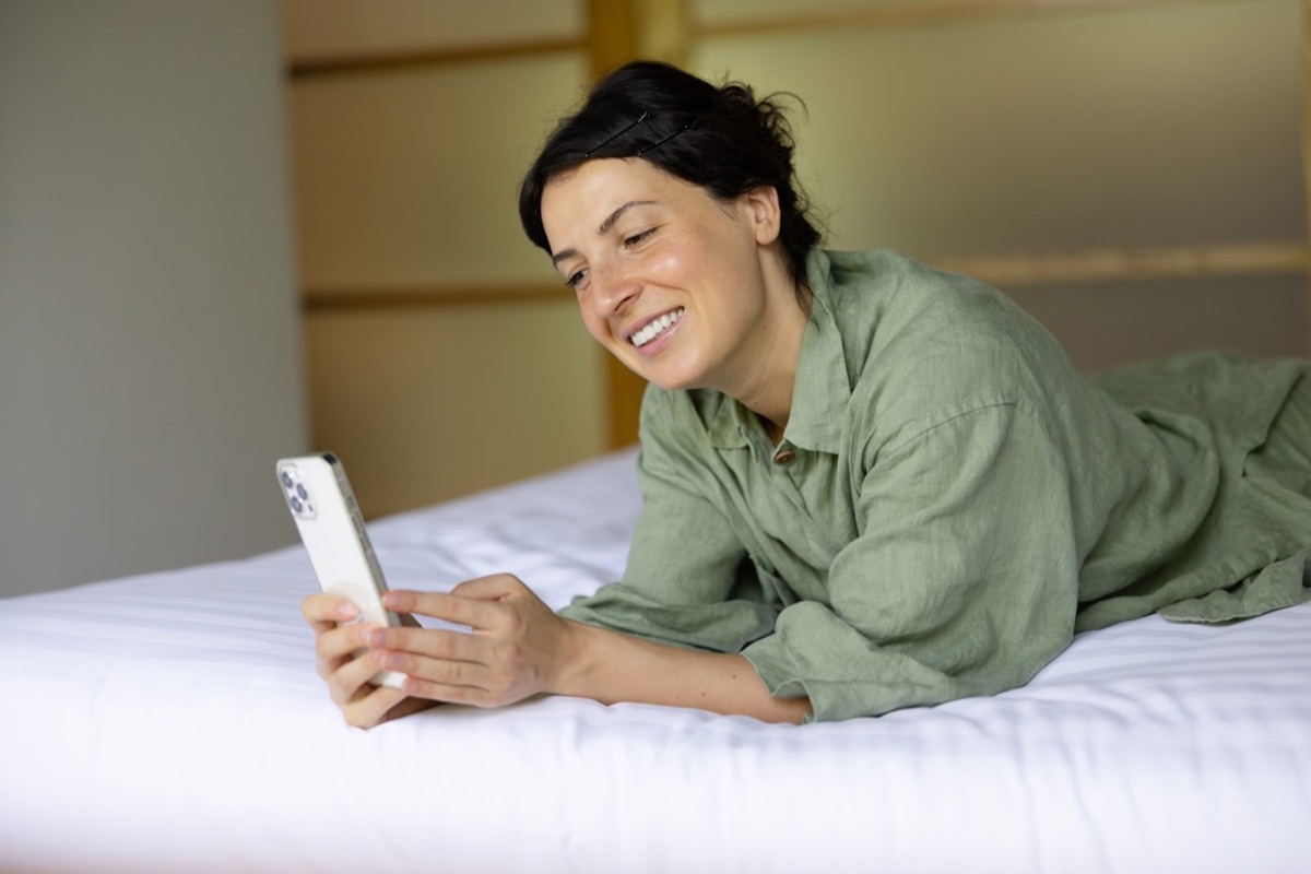 Huésped mirando el celular apoyada en la cama de un hotel
